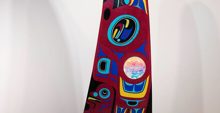 A large fin-shaped piece of glass with a red background, black top, and base made of blue metal wave shapes. The body of the "fin" is decorated with Native American designs in blue, yellow, red, black and white. A glass face is inset into the fin at lower left.