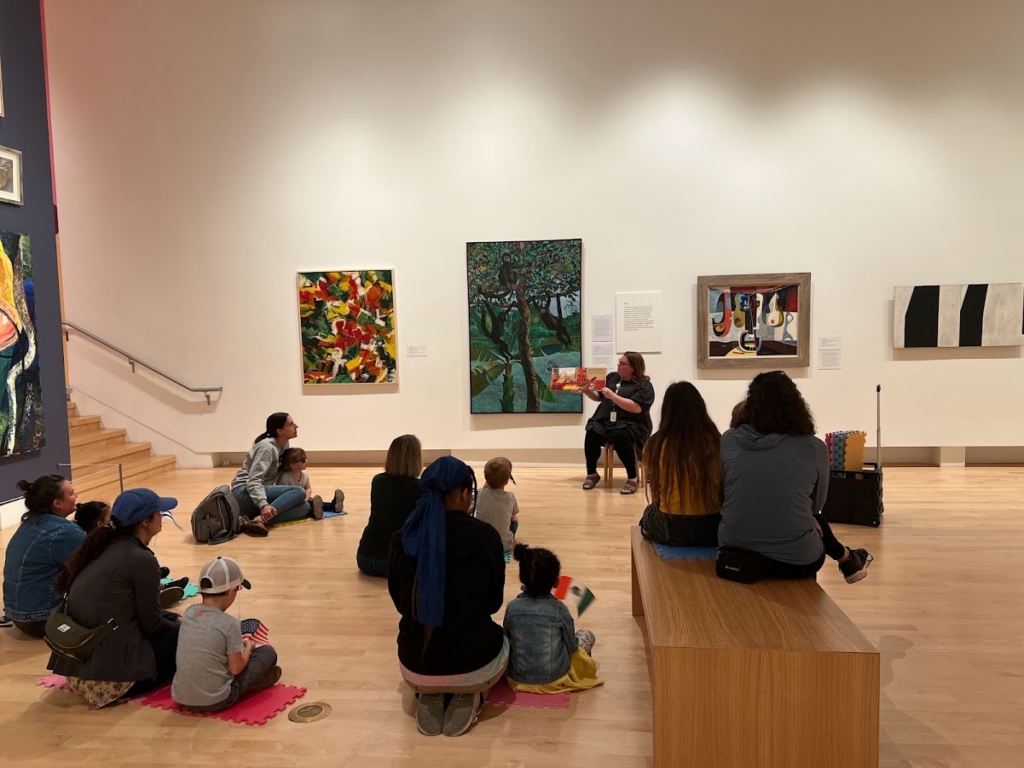 Children sitting in front of art and an educator while being read a story.